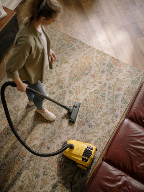 A woman using a yellow vacuum cleaner with a black hose to deep clean a floral-patterned carpet in a living room. The room features wooden flooring bordering the carpet and a red leather sofa partiall